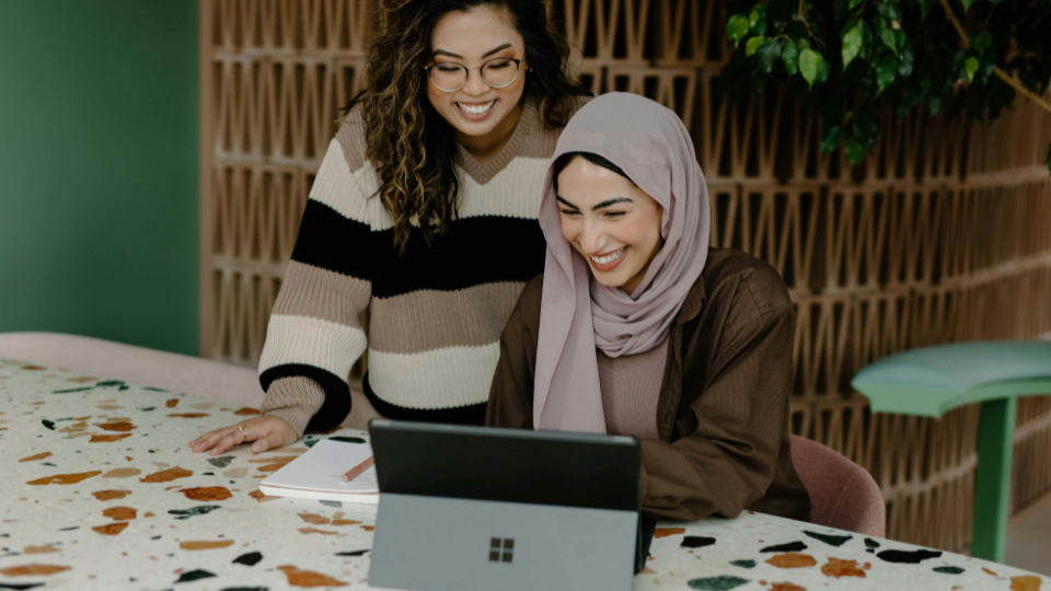Woman in a lilac hijab on a tablet, stood behind her is a woman with curly hair and a stripey jumper looking at the screen. They are both smiling